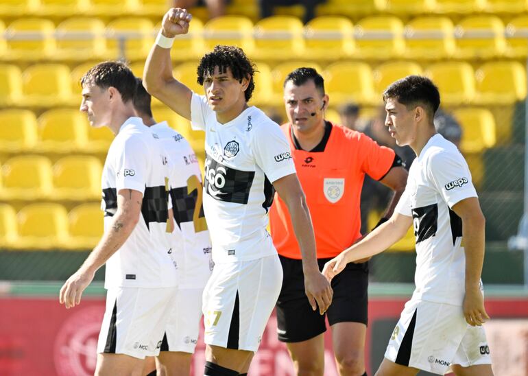 Jugadores de Olimpia con camisetas blancas levantan los brazos en celebración, mientras un árbitro supervisa al fondo en un estadio vacío.