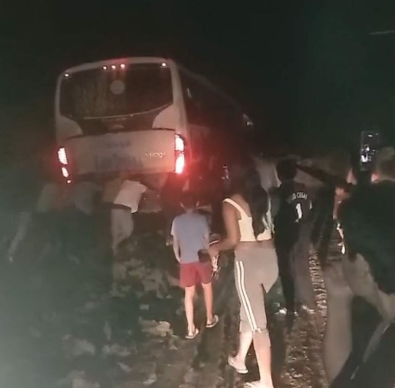 Personas empujando un autobús atascado en la carretera de tierra, rodeados de grava y con ropa casual.