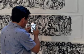 Hombre de espaldas, con camisa clara y diseños de animales, usando un teléfono móvil para capturar un mural artístico en un interior.