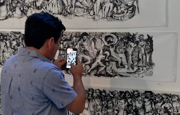 Hombre de espaldas, con camisa clara y diseños de animales, usando un teléfono móvil para capturar un mural artístico en un interior.