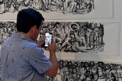 Hombre de espaldas, con camisa clara y diseños de animales, usando un teléfono móvil para capturar un mural artístico en un interior.