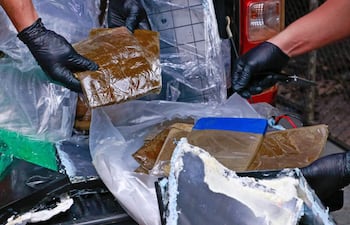 Dos personas con guantes negros manipulan paquetes envueltos en plástico en un entorno urbano.