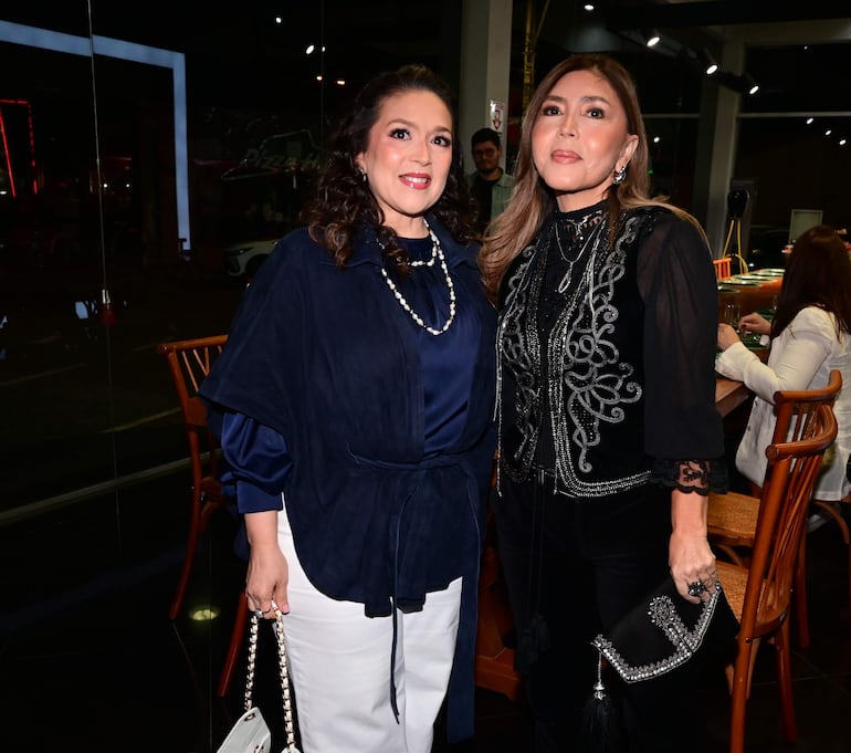 Dos mujeres sonrientes, una en blusa azul con chaqueta y otra en blusa negra, posan en un ambiente festivo.