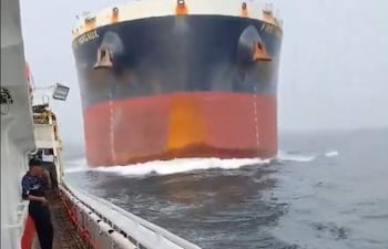Dos personas en pesquero miran hacia buque granelero azul y rojo que se aproxima en un entorno marino con niebla.