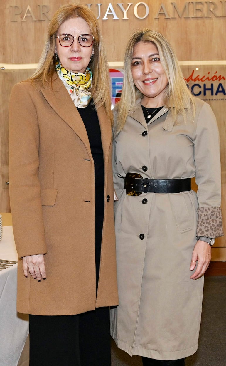 Dos mujeres sonrientes en abrigo marrón y beige, posando en un entorno formal con fondo institucional.