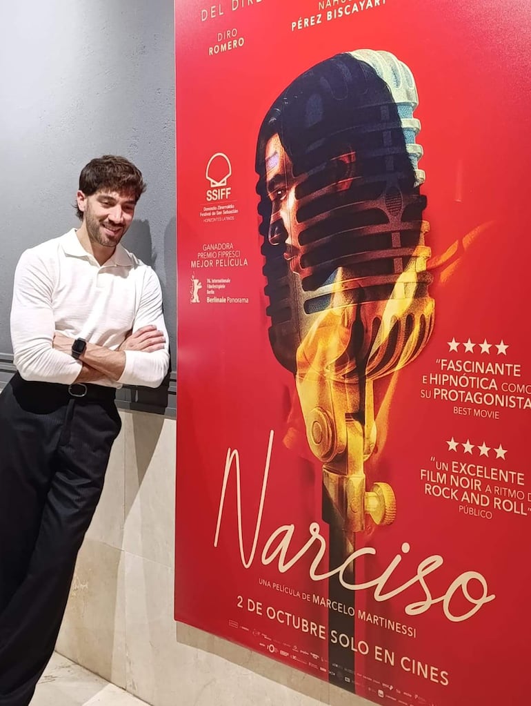 Hombre sonriendo, de cabello castaño y rizado, posando junto al cartel rojo de 'Narciso' en el evento cinematográfico.