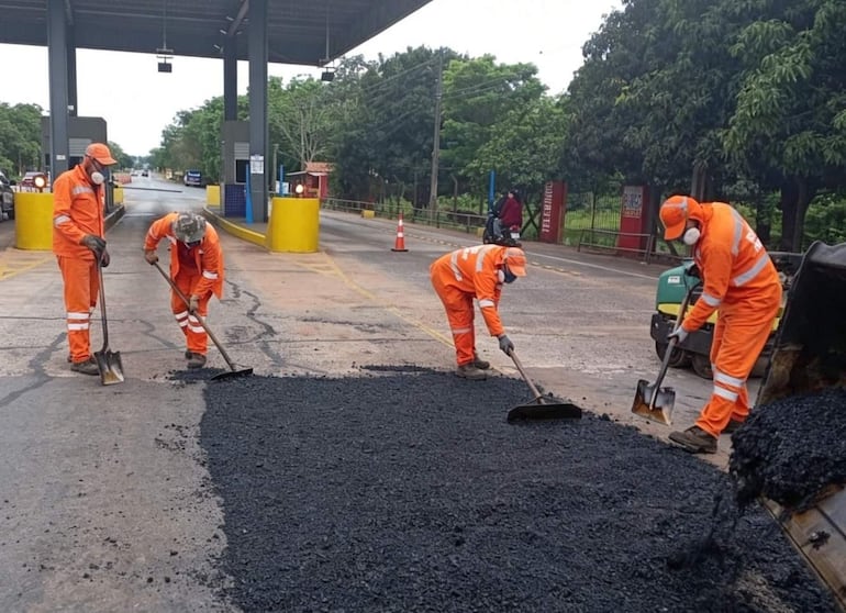 Cuatro trabajadores en trajes naranja y cascos, utilizando palas y una máquina de asfalto, en un entorno de mantenimiento vial.