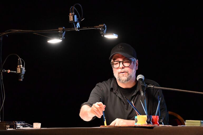 Hombre con gorra y gafas concentrado frente a un micrófono, con pinceles y pintura en un ambiente oscuro.