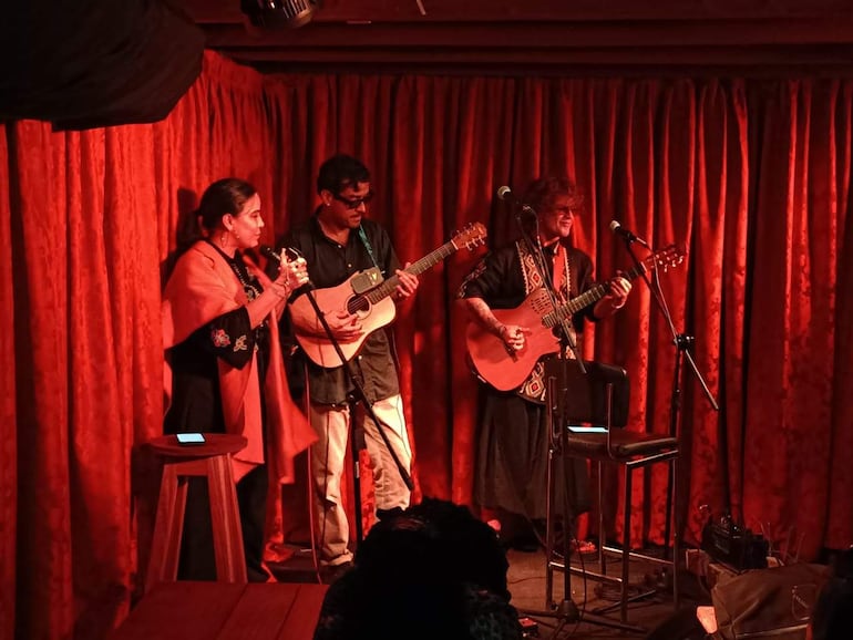 Mujer cantando con blusa bordada y chalina, mientras dos hombres tocan guitarras en un entorno íntimo y iluminado.