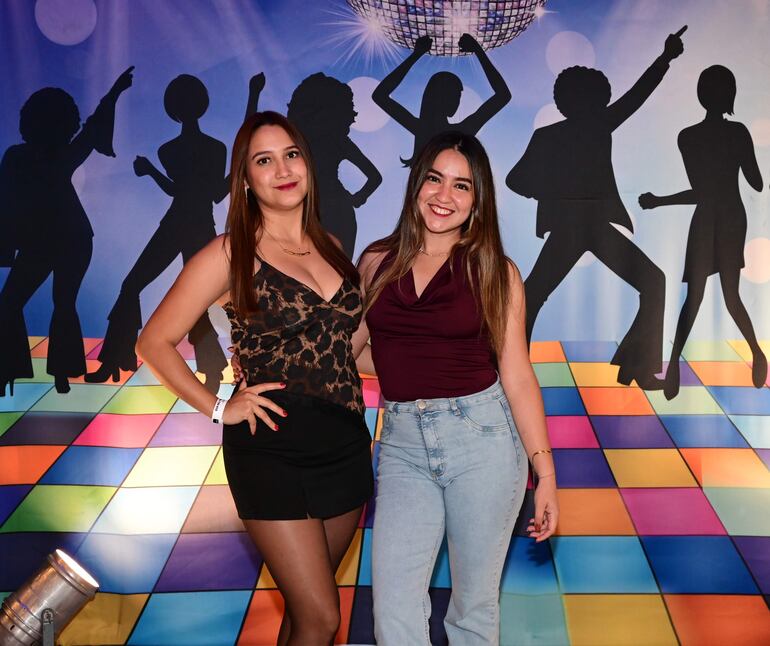 Dos mujeres sonrientes, una con blusa de estampado animal y la otra con blusa burdeos, posan en un fondo colorido en un evento festivo.