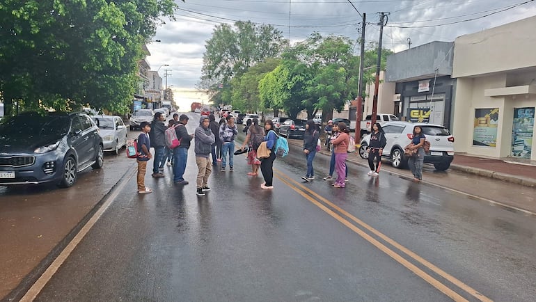 Grupo de indígenas conversando en la calle, algunos llevan mochilas, con comercios locales de fondo y calles mojadas por la lluvia.