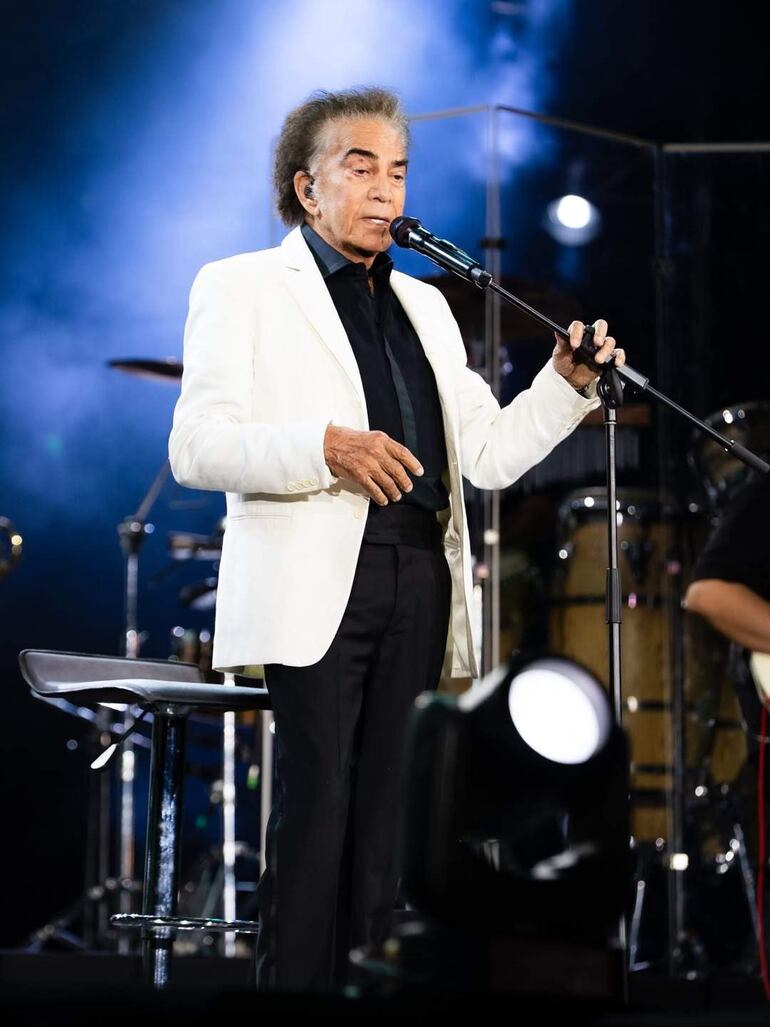 Hombre mayor, cabello rizado y canoso, vestido con traje blanco, canta en el escenario con ambiente oscuro y luces de concierto.