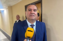 Derlis Maidana, vestido de traje azul y corbata, sonríe mientras sostiene un micrófono amarillo. Su asistente en segundo plano tiene una expresión seria.