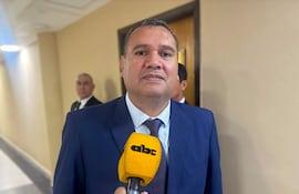 Derlis Maidana, vestido de traje azul y corbata, sonríe mientras sostiene un micrófono amarillo. Su asistente en segundo plano tiene una expresión seria.