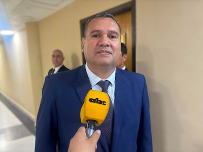 Derlis Maidana, vestido de traje azul y corbata, sonríe mientras sostiene un micrófono amarillo. Su asistente en segundo plano tiene una expresión seria.