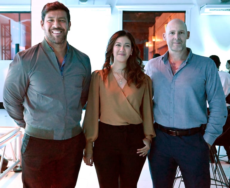 Hombre con chaqueta ligera, mujer con blusa marrón y hombre con camisa azul, todos sonriendo en un ambiente moderno.