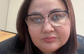 Mujer con cabello oscuro y largo, blusa negra y gafas, miran a la cámara en un entorno de oficina neutro.