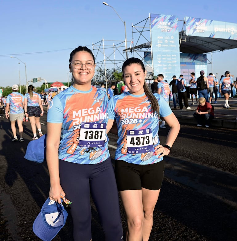 Dos mujeres, una con camiseta azul y gafas, sostienen gorras mientras sonríen en un ambiente deportivo al aire libre.