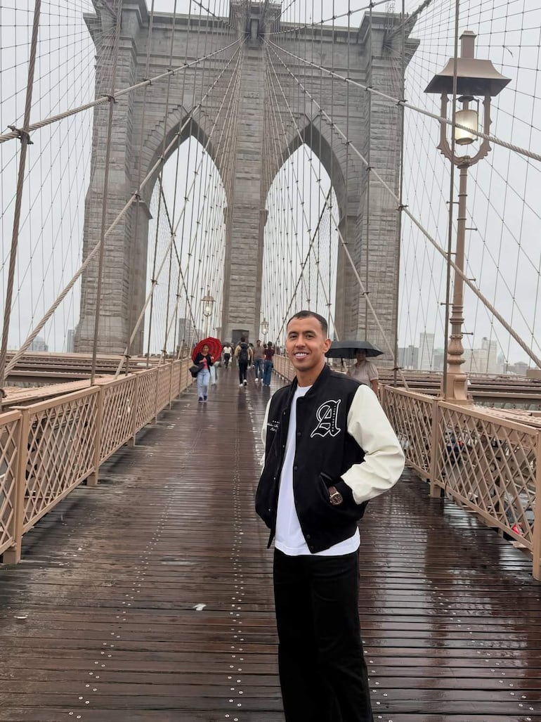 Alejandro Kaku Romero sonríe con chaqueta negra y camiseta blanca, rodeado de personas con paraguas en el Puente de Brooklyn.