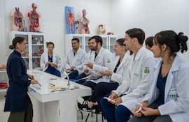 Instructora de pie con puntero rodeada de estudiantes en batas blancas, todos en aula con modelos anatómicos y materiales educativos.