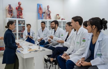 Instructora de pie con puntero rodeada de estudiantes en batas blancas, todos en aula con modelos anatómicos y materiales educativos.