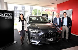 Mujer sonriente con traje blanco y negro junto al vehículo gris Honda ZR-V, acompañada de dos hombres en un ambiente moderno y profesional.