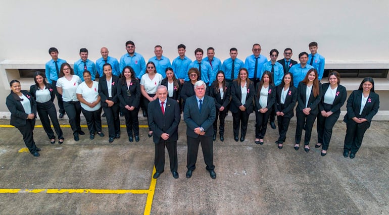 Dos hombres con trajes elegantes al centro de un grupo de 30 personas, todos con expresiones felices, en un ambiente exterior.