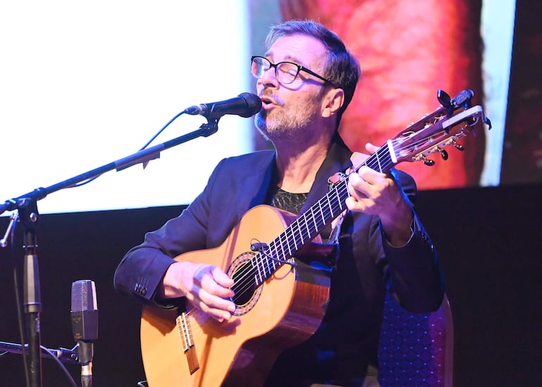 Kevin Johansen, con anteojos y saco oscuro, toca la guitarra en un escenario iluminado, concentrado en su actuación.