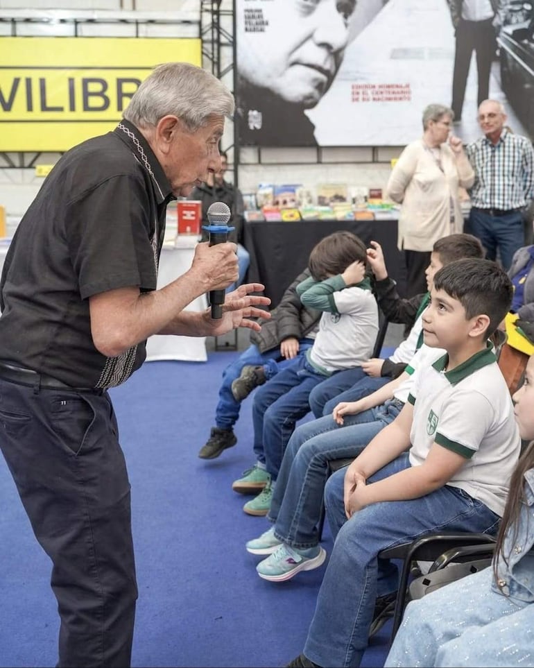 Hombre mayor con micrófono habla a niños sentados, en feria de libros con estantes y cartel de 'VILIBRIO'.
