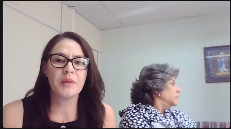 Rocío Vega con blusa negra y gafas, habla al público mientras otra mujer de cabello canoso escucha pensativa.