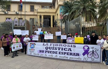 Grupo de personas con camisetas púrpuras sostiene pancartas en protesta frente a un edificio gubernamental. Baner visible dice 'LA FIBROSIS QUÍSTICA NO PUEDE ESPERAR'.