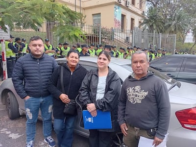 Hombre con abrigo azul y mujer con abrigo negro sostienen documentos mientras son fotografiados en un ambiente exterior.