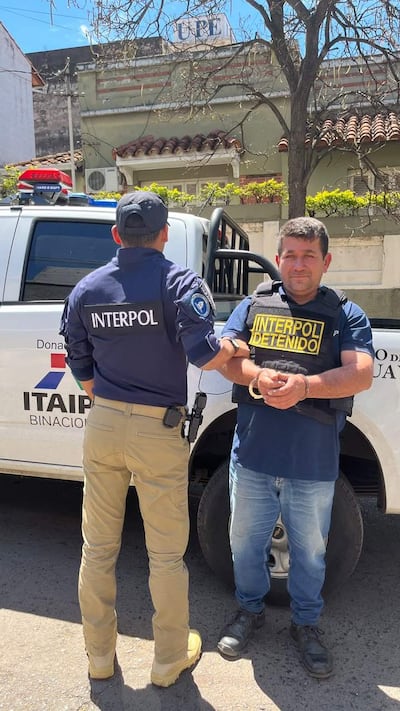 Hombre esposado con chaleco de INTERPOL y agente uniformado detrás en un operativo al aire libre.