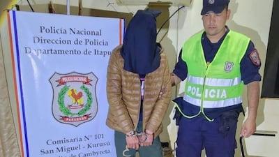 Individuo con capuchón negro y chaqueta marrón, esposado, junto a un policía en uniforme amarillo en un ambiente interior.