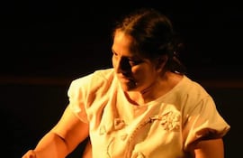 Mujer con vestimenta clara, enfocada en un objeto de tejido, en un entorno oscuro que sugiere un escenario teatral.