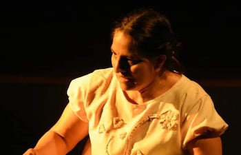Mujer con vestimenta clara, enfocada en un objeto de tejido, en un entorno oscuro que sugiere un escenario teatral.