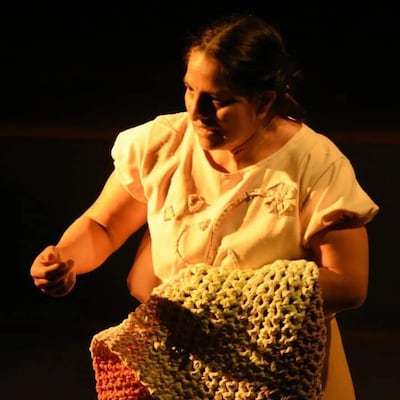 Mujer con vestimenta clara, enfocada en un objeto de tejido, en un entorno oscuro que sugiere un escenario teatral.