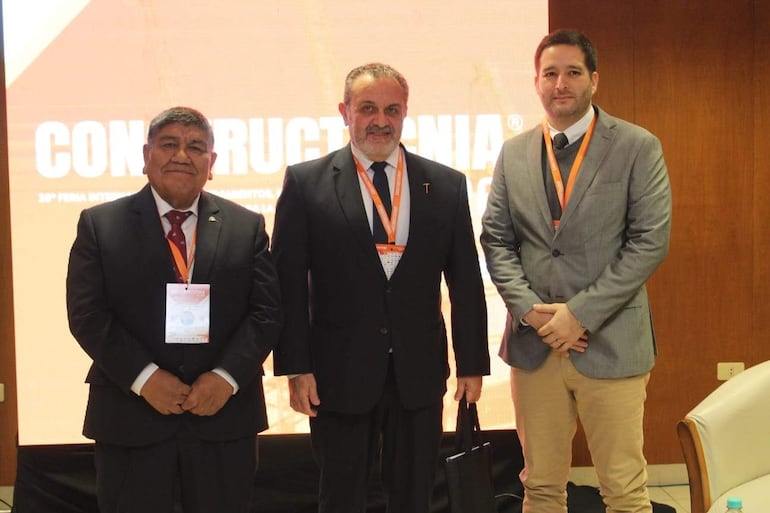 Tres hombres de pie en trajes formales discuten en un evento de minería, con credenciales visibles y una pantalla detrás.