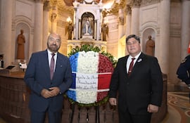 Pierre-Christian Soccoja sonríe en traje azul y corbata a rayas, mientras otro hombre en traje negro con corbata roja lo acompaña ante el altar decorado.