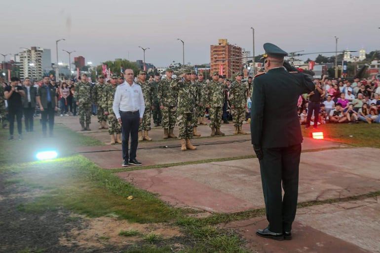 Grupo de soldados en uniforme camuflado con un militar saludando, fondo urbano con espectadores atentos al evento.