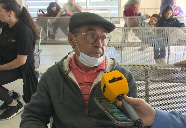 Hombre mayor con gorra y gafas, camiseta rosa y abrigo gris, habla con un periodista en sala de espera.