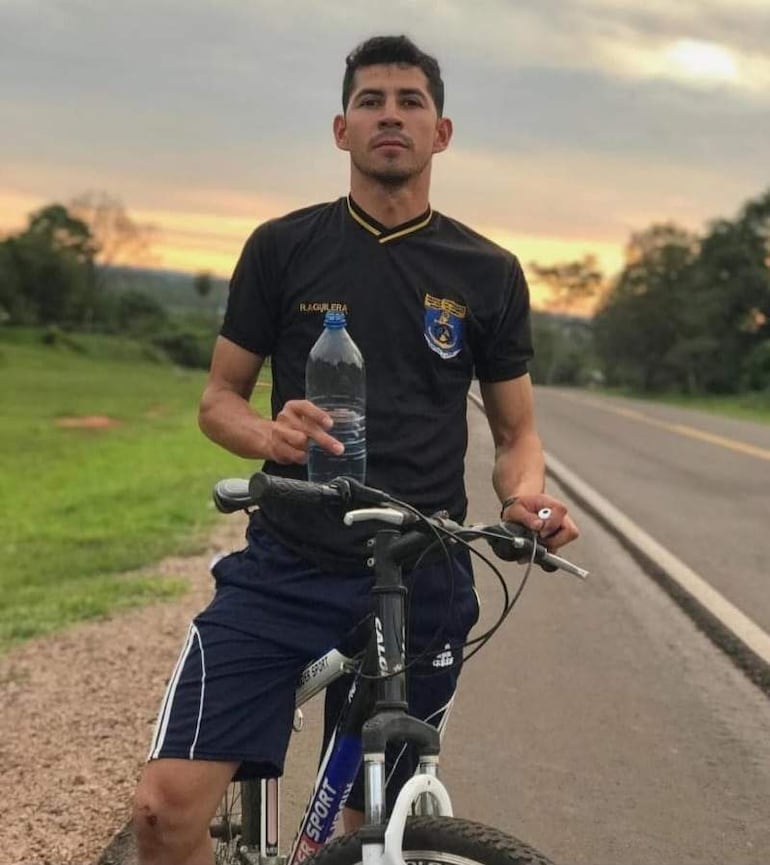 Joven Ricardo Aguilera León de camiseta negra y pantalones cortos, sostiene una botella de agua junto a su bicicleta en una carretera.