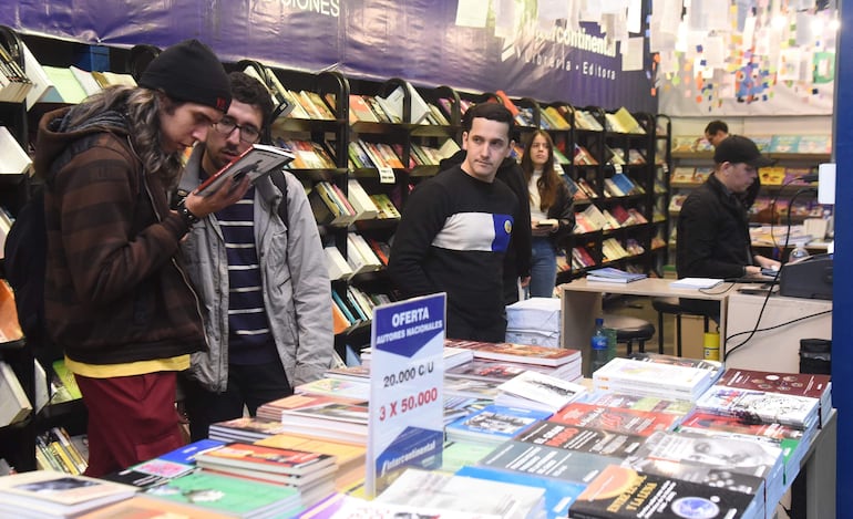 Hombre con cabello largo revisa un libro, otro con gafas observa, mientras una mujer y un hombre con computadora están presentes.