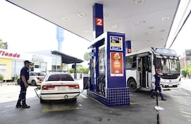 Empleado de estación de servicio en uniforme azul abastece un automóvil mientras un bus de Policía está presente.