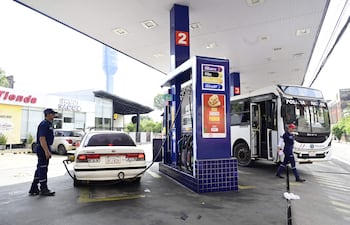 Empleado de estación de servicio en uniforme azul abastece un automóvil mientras un bus de Policía está presente.