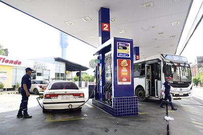 Empleado de estación de servicio en uniforme azul abastece un automóvil mientras un bus de Policía está presente.
