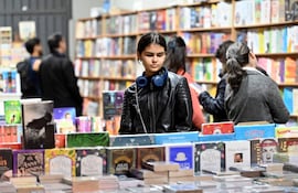 Joven de cabello oscuro con chaqueta de cuero y auriculares, mirando libros en una mesa repleta en ambiente de feria literaria.