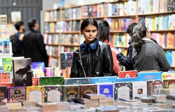 Joven de cabello oscuro con chaqueta de cuero y auriculares, mirando libros en una mesa repleta en ambiente de feria literaria.
