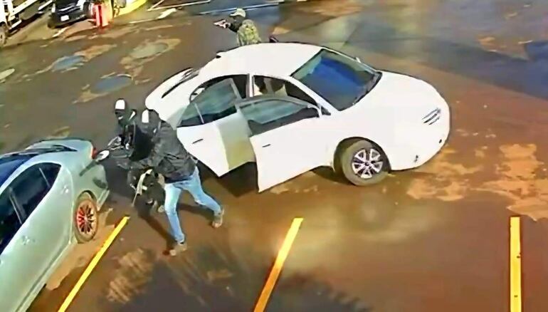 Dos individuos encapuchados con vestimenta oscura manipulando una motocicleta cerca de un vehículo en un estacionamiento.
