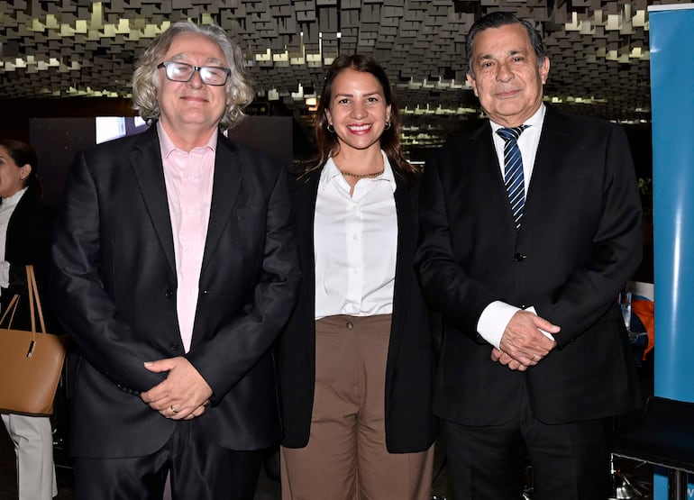 Hombres y mujer conversan, el primero con saco negro y camisa rosa, la mujer en blusa blanca y pantalón marrón, el tercero con traje negro y corbata azul.